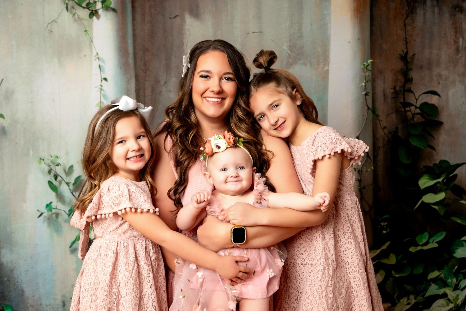A smiling mother in pink poses with three young daughters, all wearing matching pink dresses, in a beautifully styled portrait.