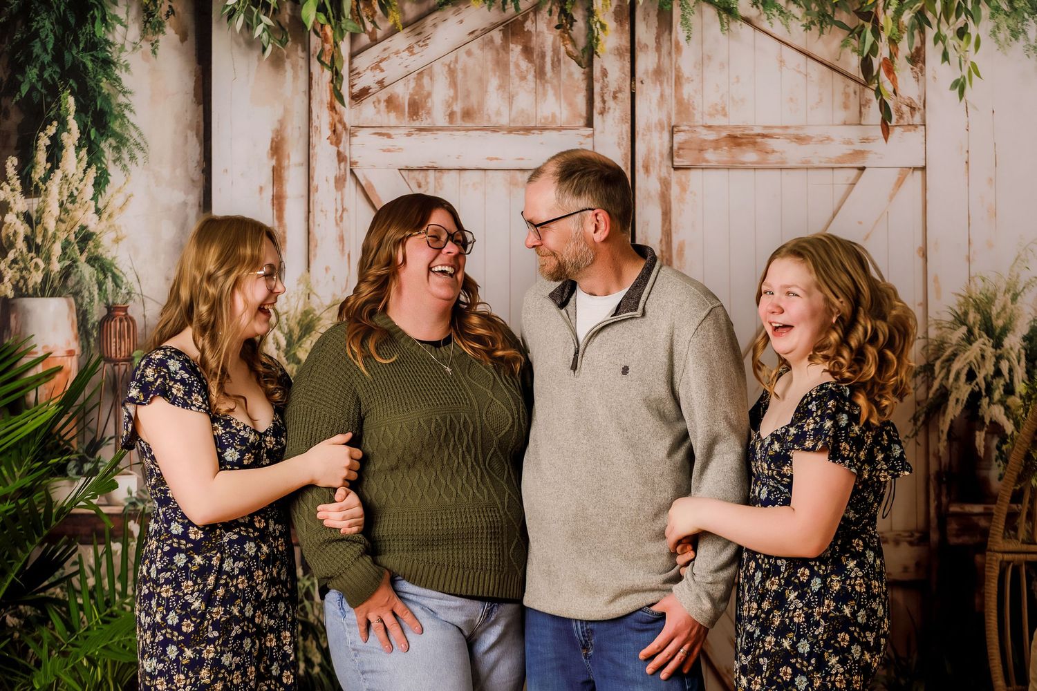 Family of four laughing together in front of rustic barn doors surrounded by greenery in a warm portrait session.