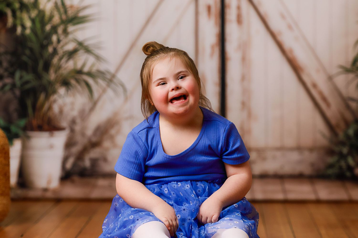 Joyful young girl with Down syndrome in a blue dress sitting on a wooden floor, smiling brightly indoors.