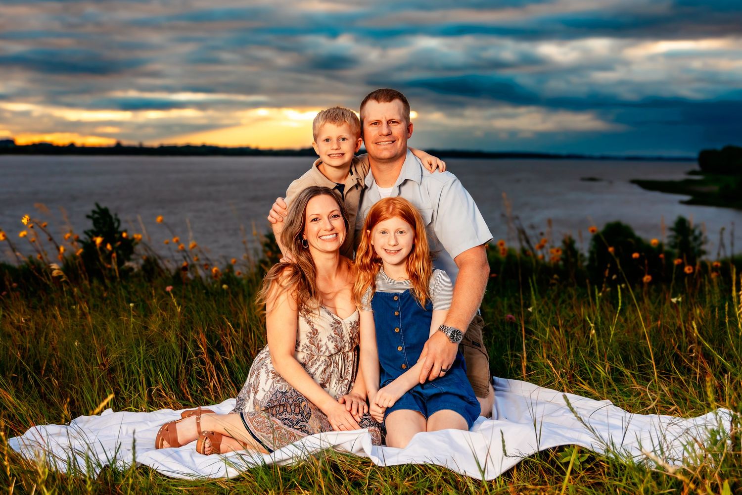 A rustic outdoor family photoshoot featuring scenic lakeside views at sunset with natural grass and dramatic clouds.