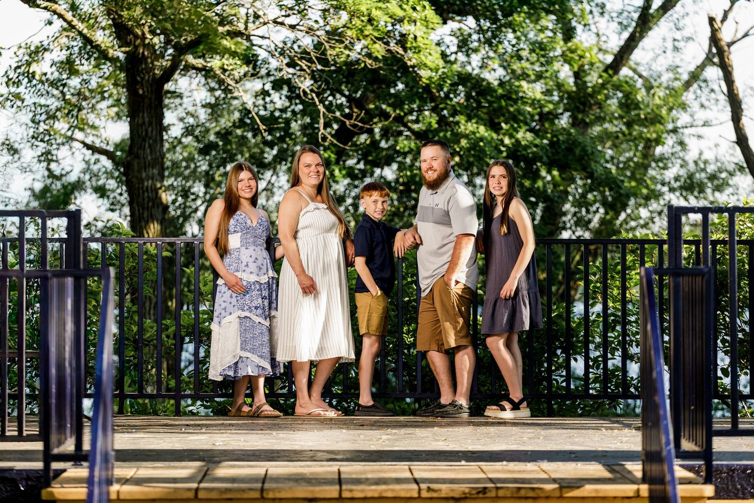 Family of five posing outdoors on a deck with lush green trees and a black iron fence backdrop.