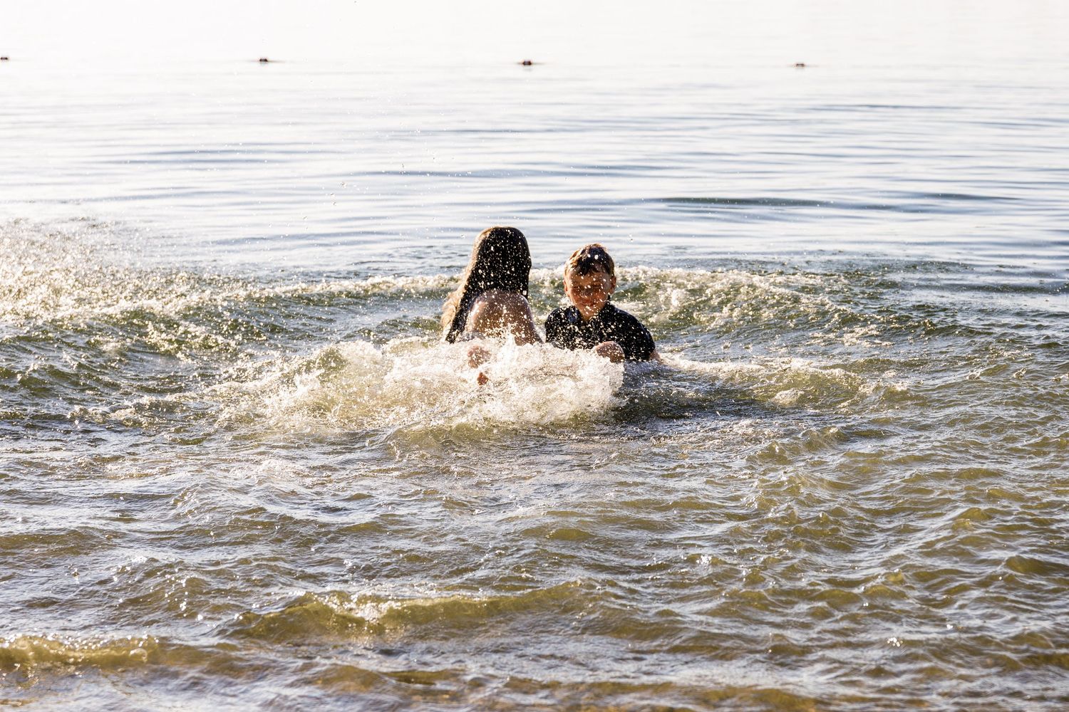 Two children laughing and swimming together in open lake water on a bright sunny day.