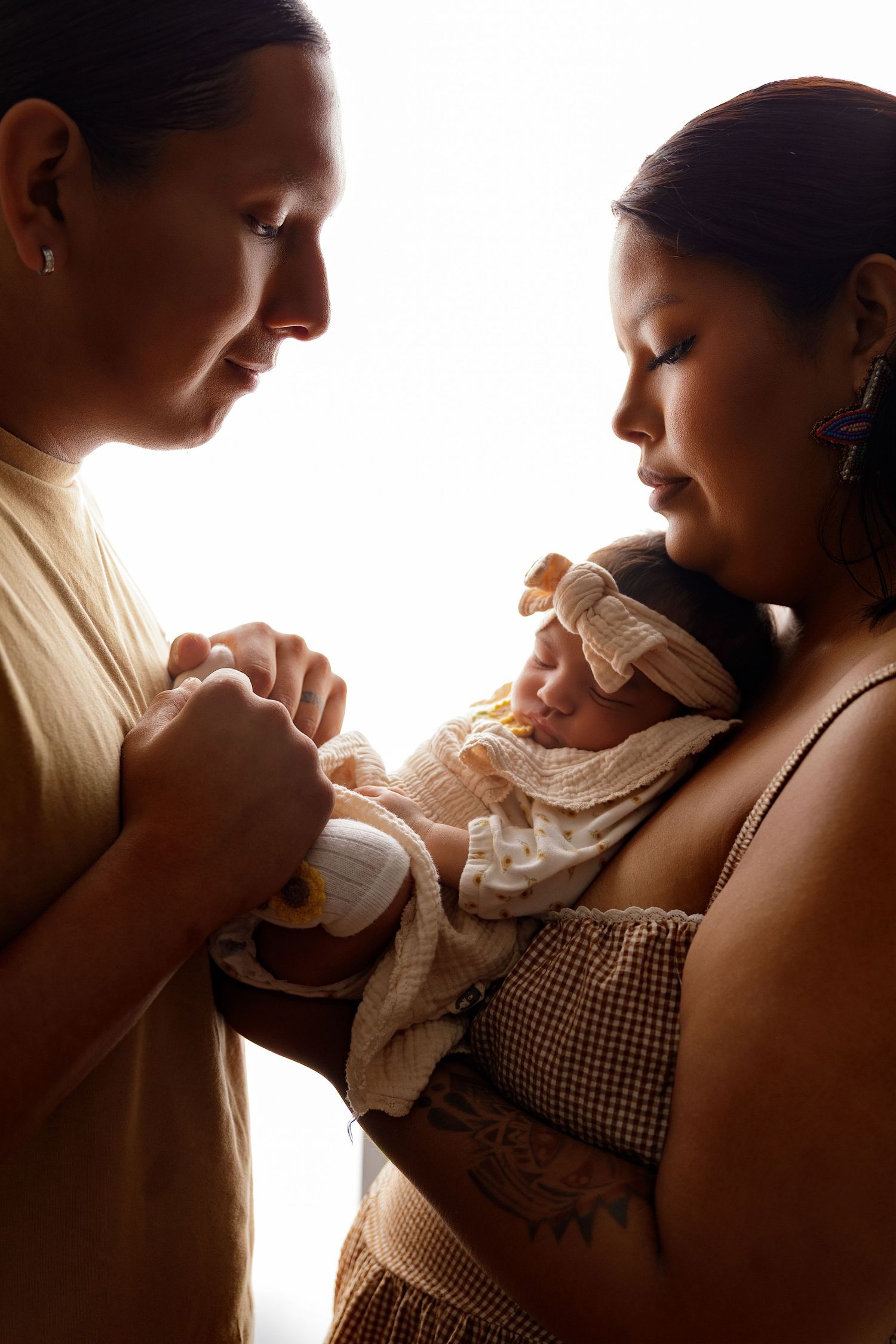 Two mothers tenderly holding their newborn baby wrapped in a white blanket with a bow headband in warm backlit light.