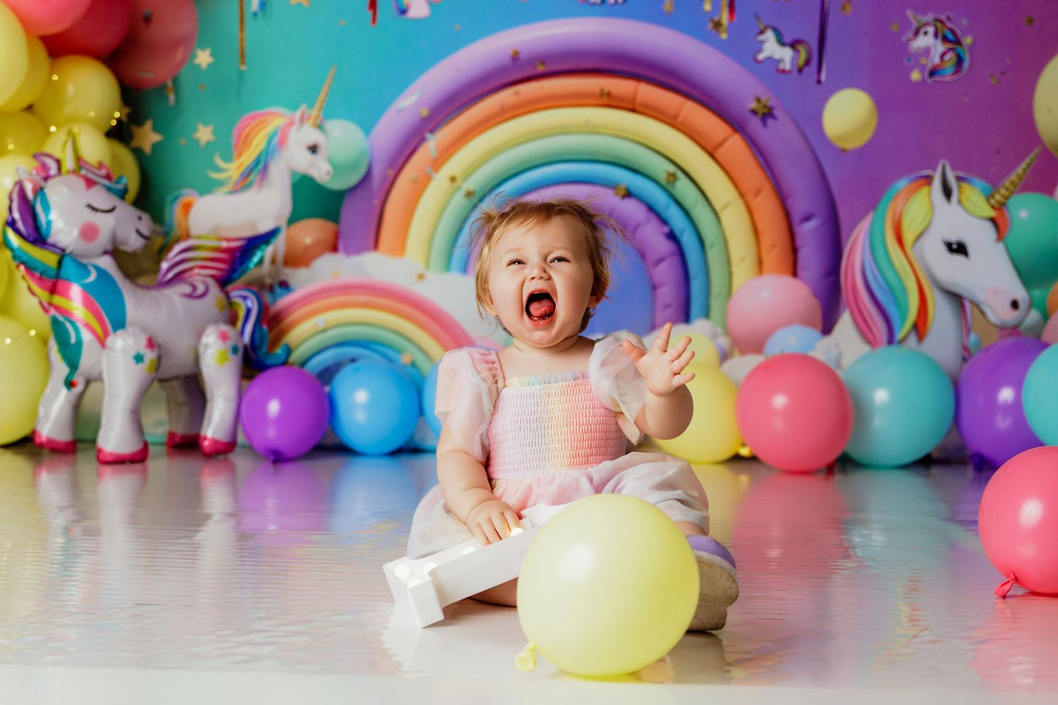 Baby girl laughing at unicorn-themed first birthday party with colorful balloons and rainbow decorations.