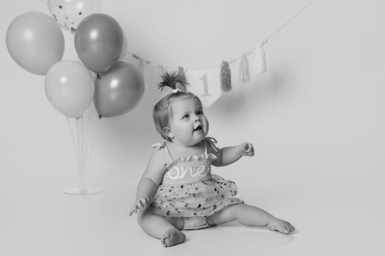 Baby sitting by grey balloons and clothesline in floral romper.
