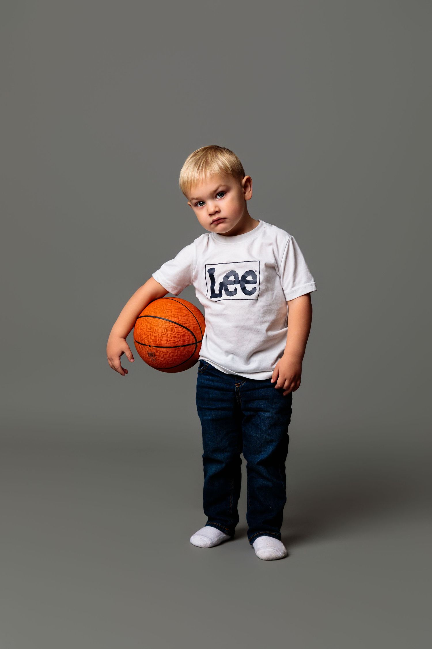 A young child in a white t-shirt and jeans holds a basketball while posing against a gray studio backdrop.