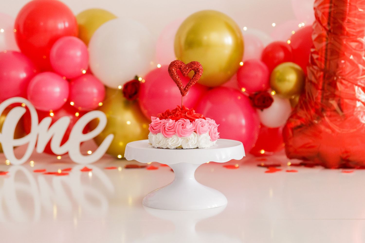 Pink, red, and gold balloon garland decorates a white wall with festive party decorations.