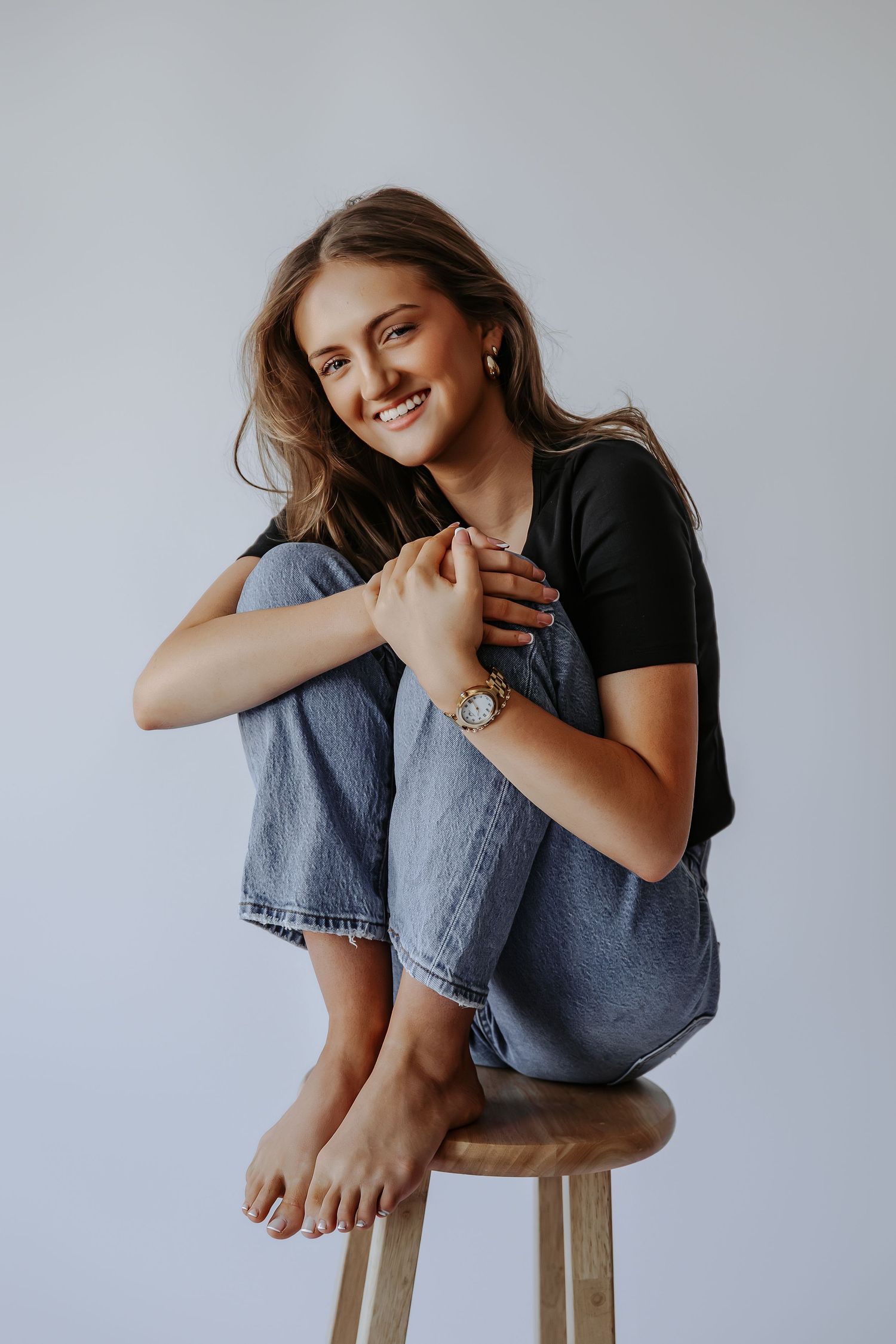 Smiling young woman in black tee and blue jeans sitting barefoot on wooden stool in studio setting.