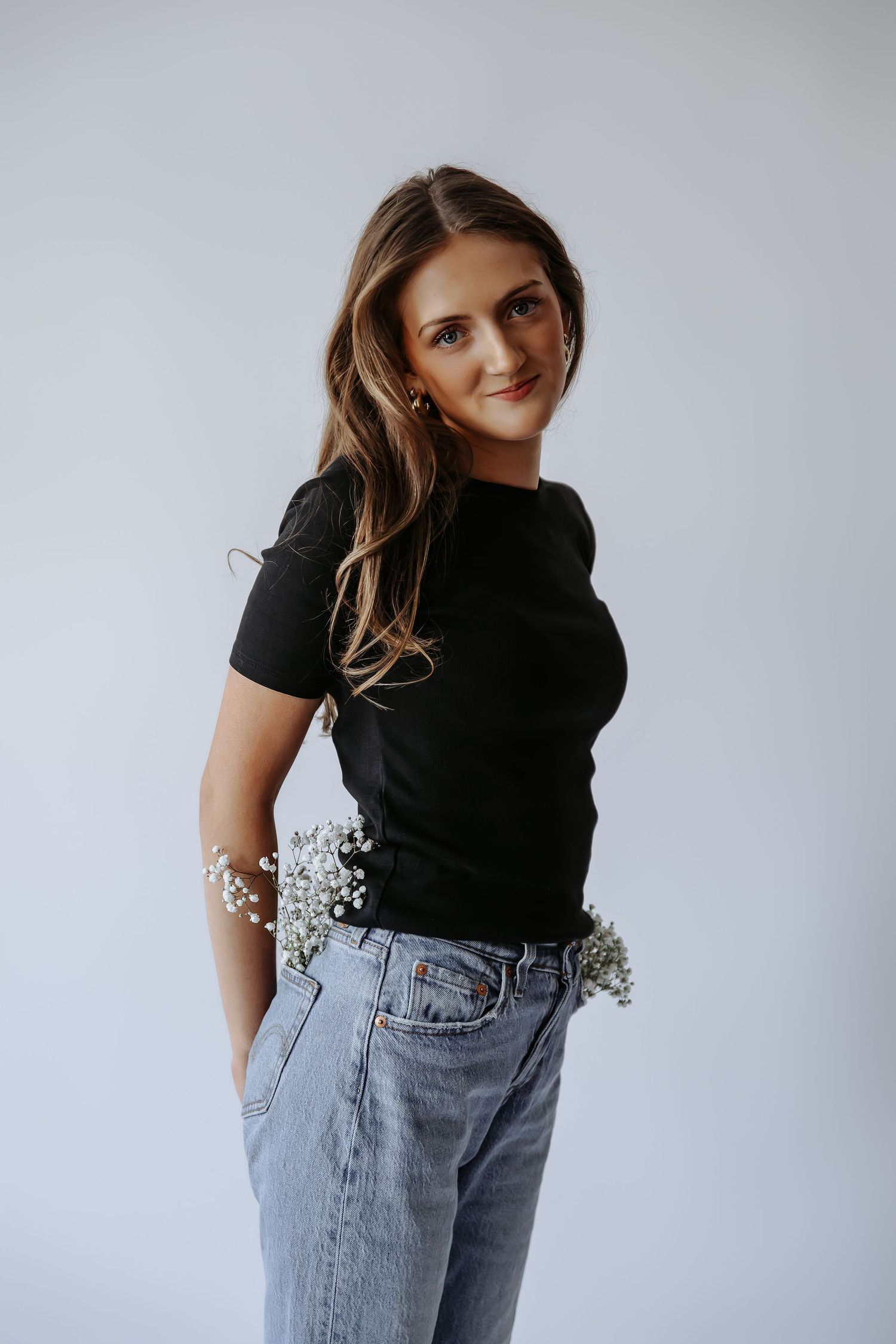 Young woman in black fitted tee and light jeans, smiling over shoulder, white baby's breath flowers tucked in waistband, gray background.