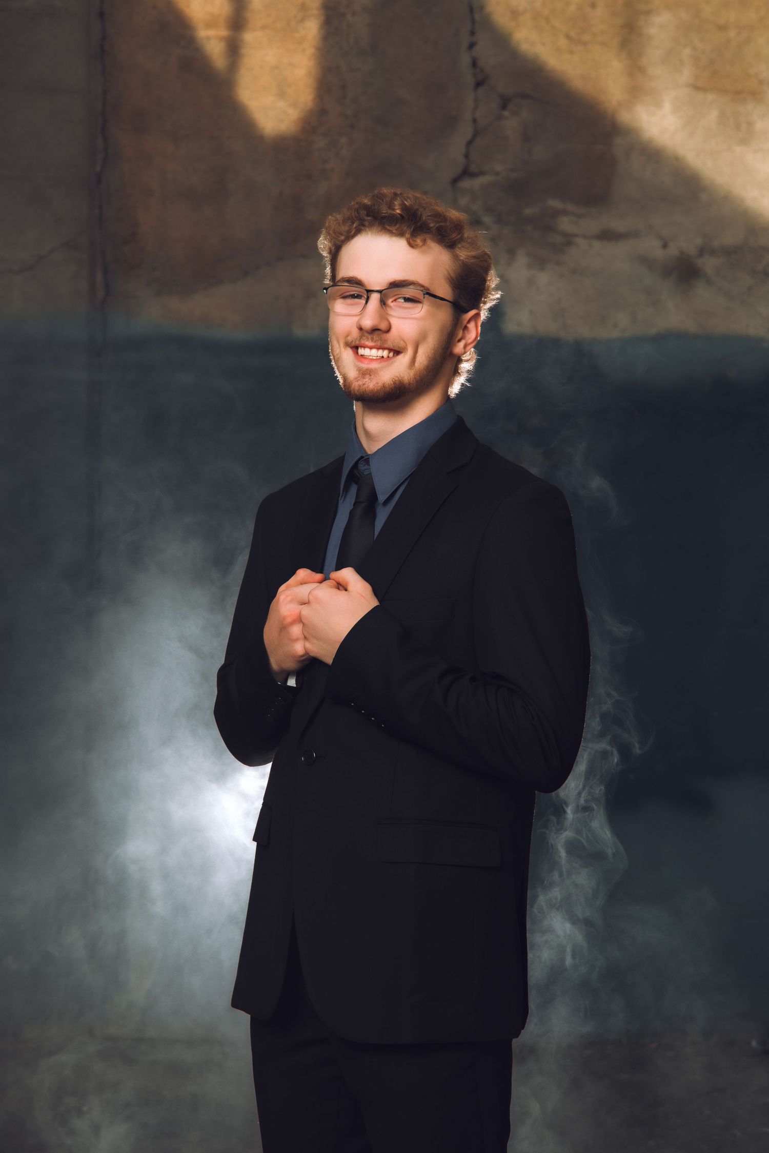 Young man in black suit and tie with glasses smiling confidently in a smoky studio setting.
