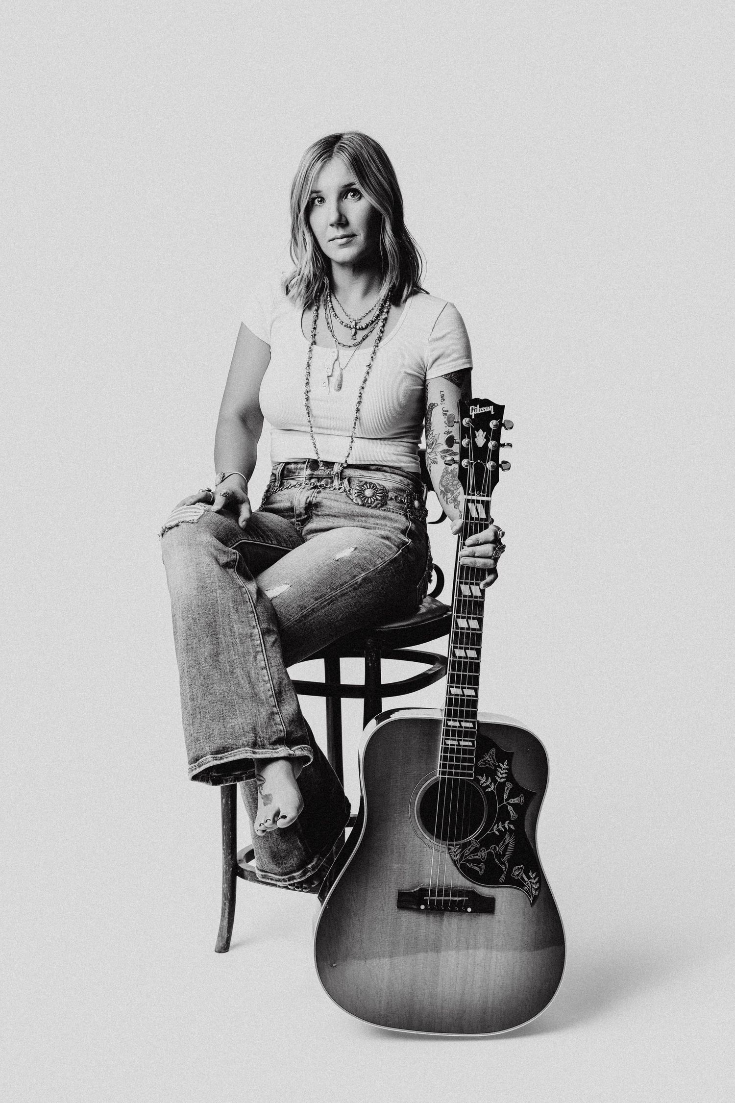 Black and white photo of a woman sitting on a stool holding an acoustic guitar, wearing jeans and a white t-shirt.