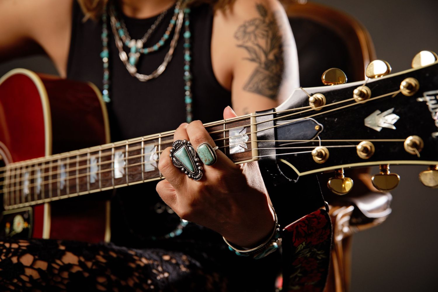 Close-up of tattooed hands playing acoustic guitar with turquoise rings and bracelets.