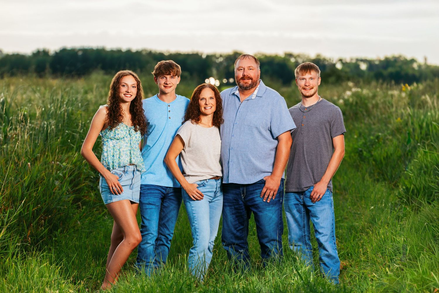 Family wearing coordinated blue and white outfits poses together in tall grass field during golden hour.