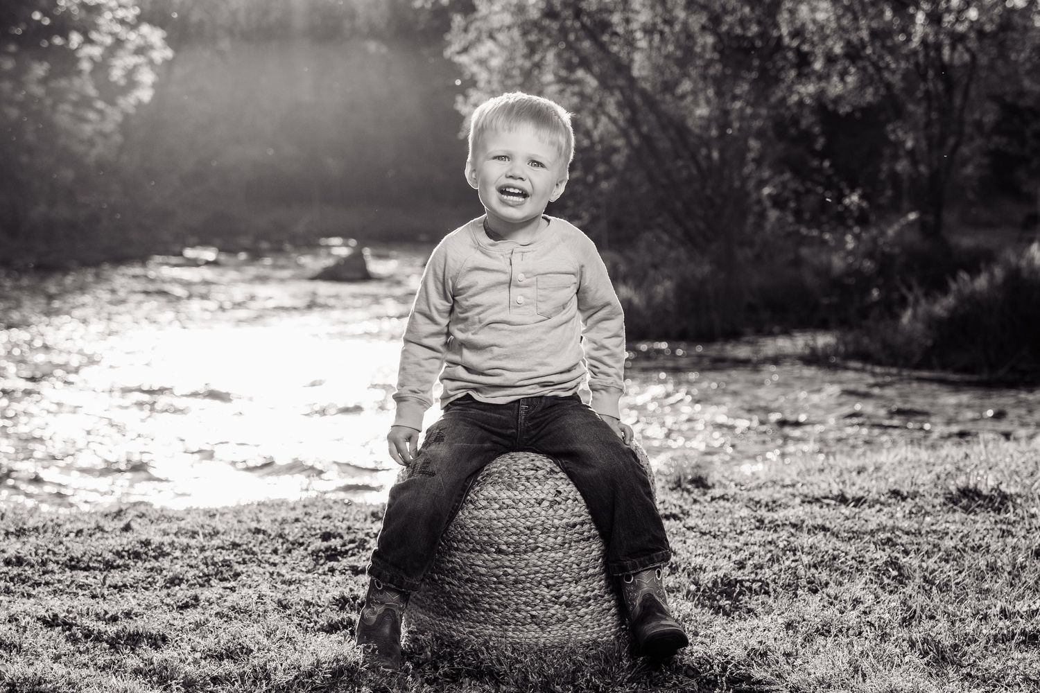 Black and white photo of a laughing toddler boy sitting on a rock outdoors near a sunlit stream.