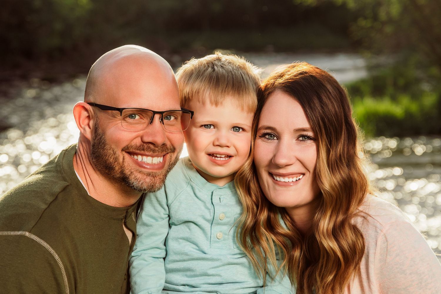 Happy family portrait of smiling parents with young toddler boy outdoors near sparkling water in warm sunlight.