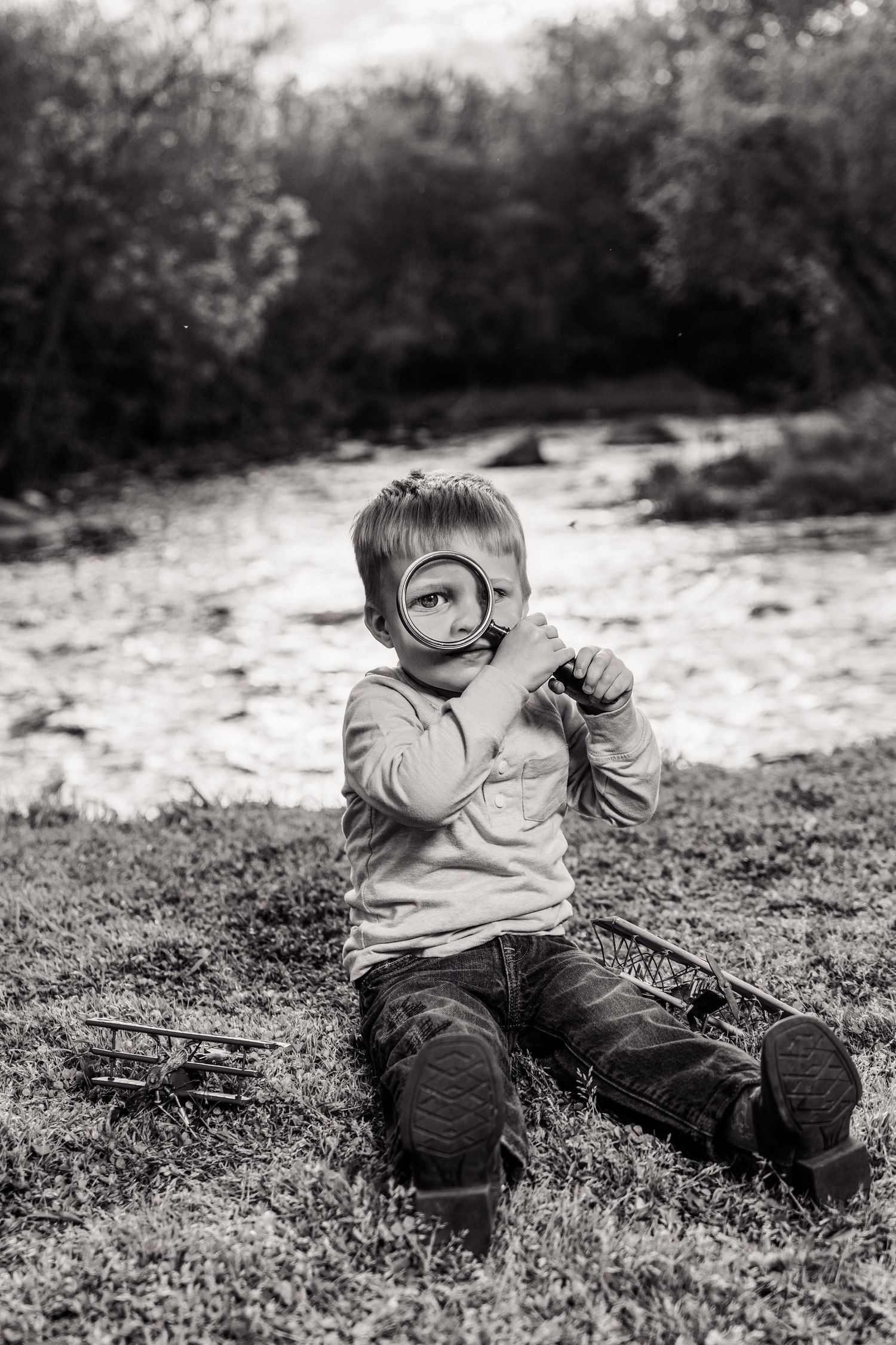 Black and white photo of a young boy sitting outdoors holding a magnifying glass up to his eye near a river.