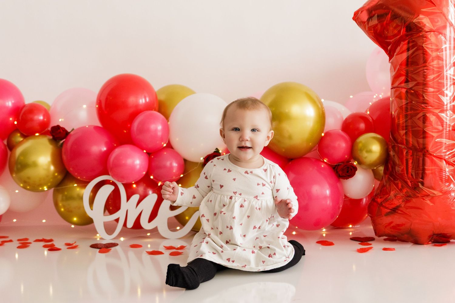 Festive first birthday photo setup with red and gold balloons and decorative 'one' sign.