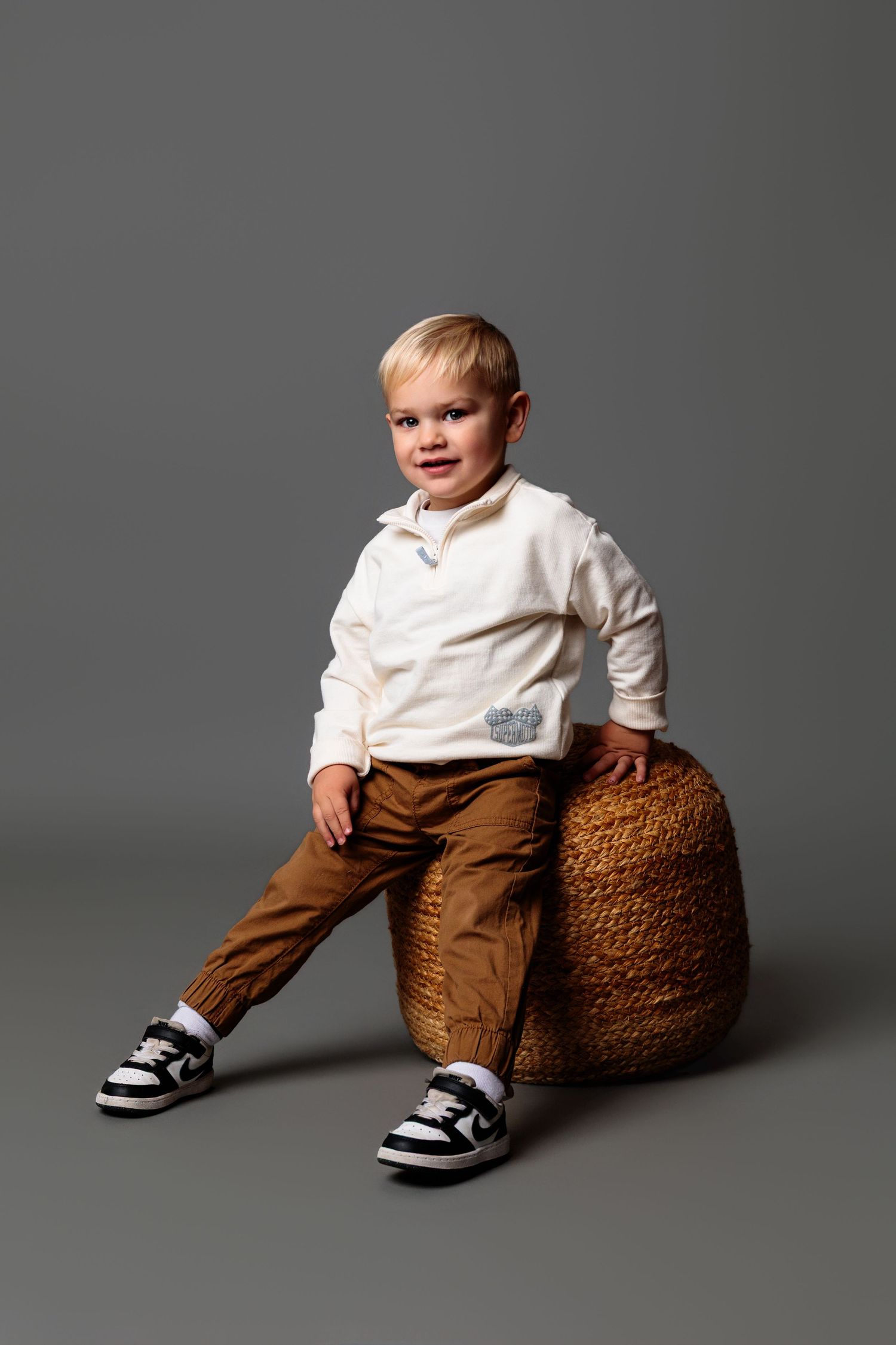 Small child sits on a brown pouf wearing brown pants and black sneakers in a studio setting.