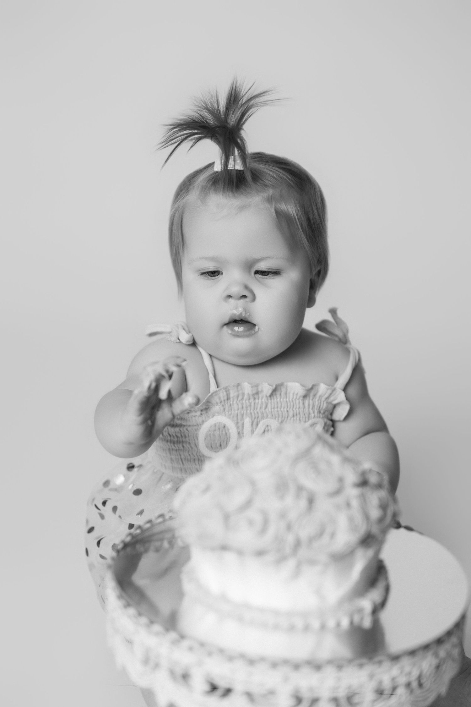 Baby in ruffled outfit explores white frosted cake during smash cake session.
