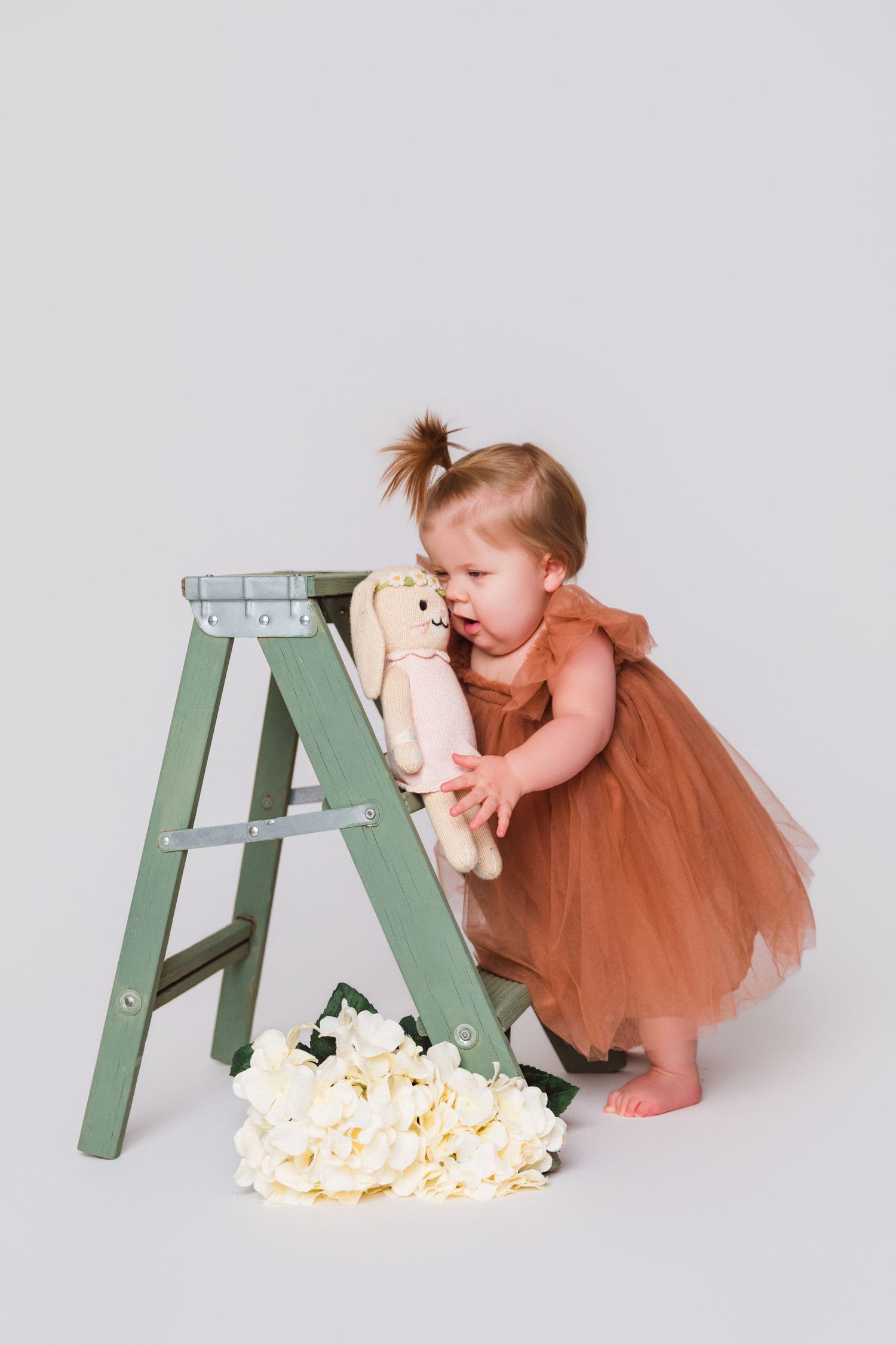 A toddler in an orange tulle dress hugs a stuffed animal while standing near a green ladder.