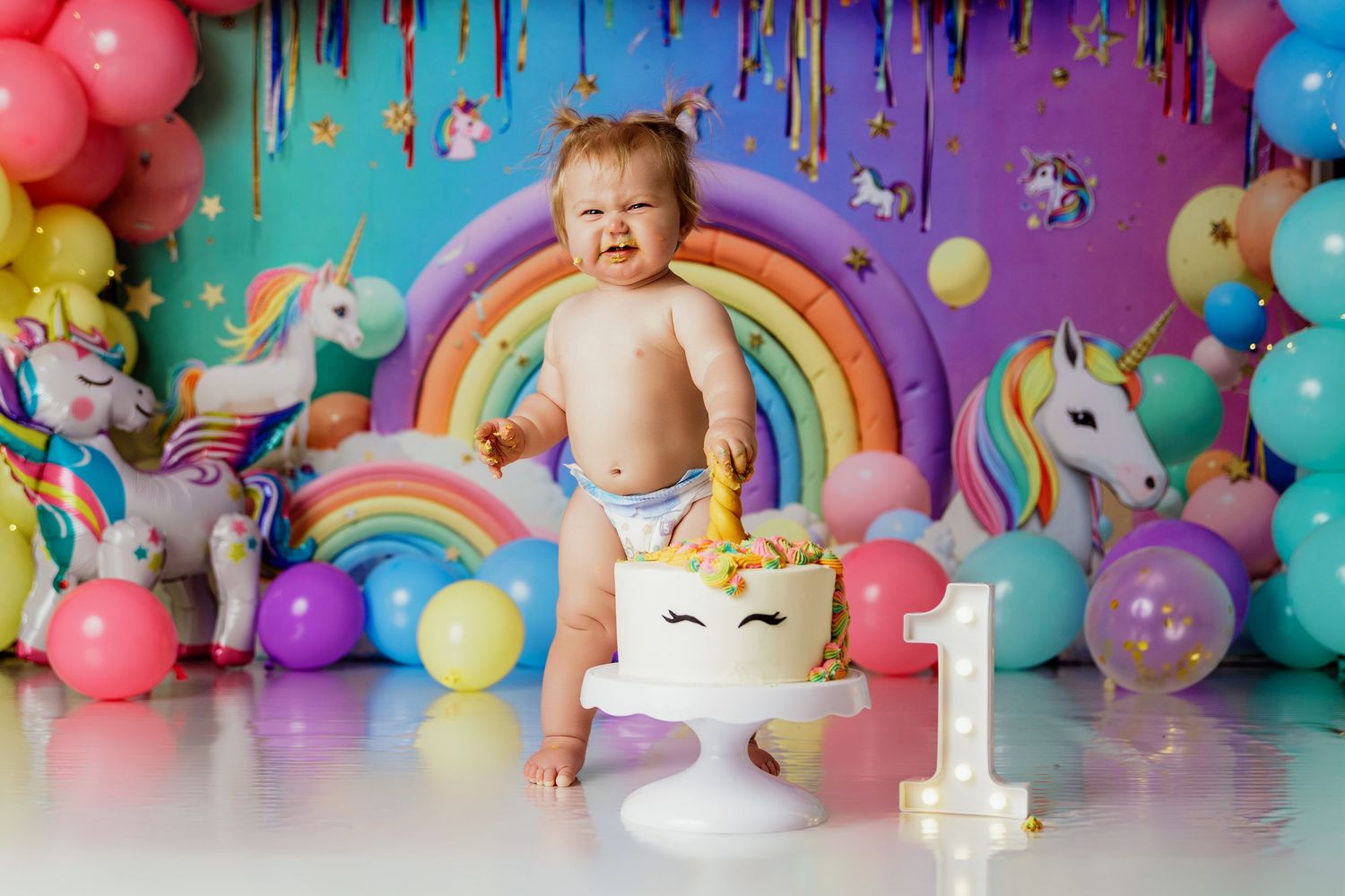 Baby's first birthday cake smash with unicorn theme, colorful balloons, rainbow backdrop, and number one decoration.