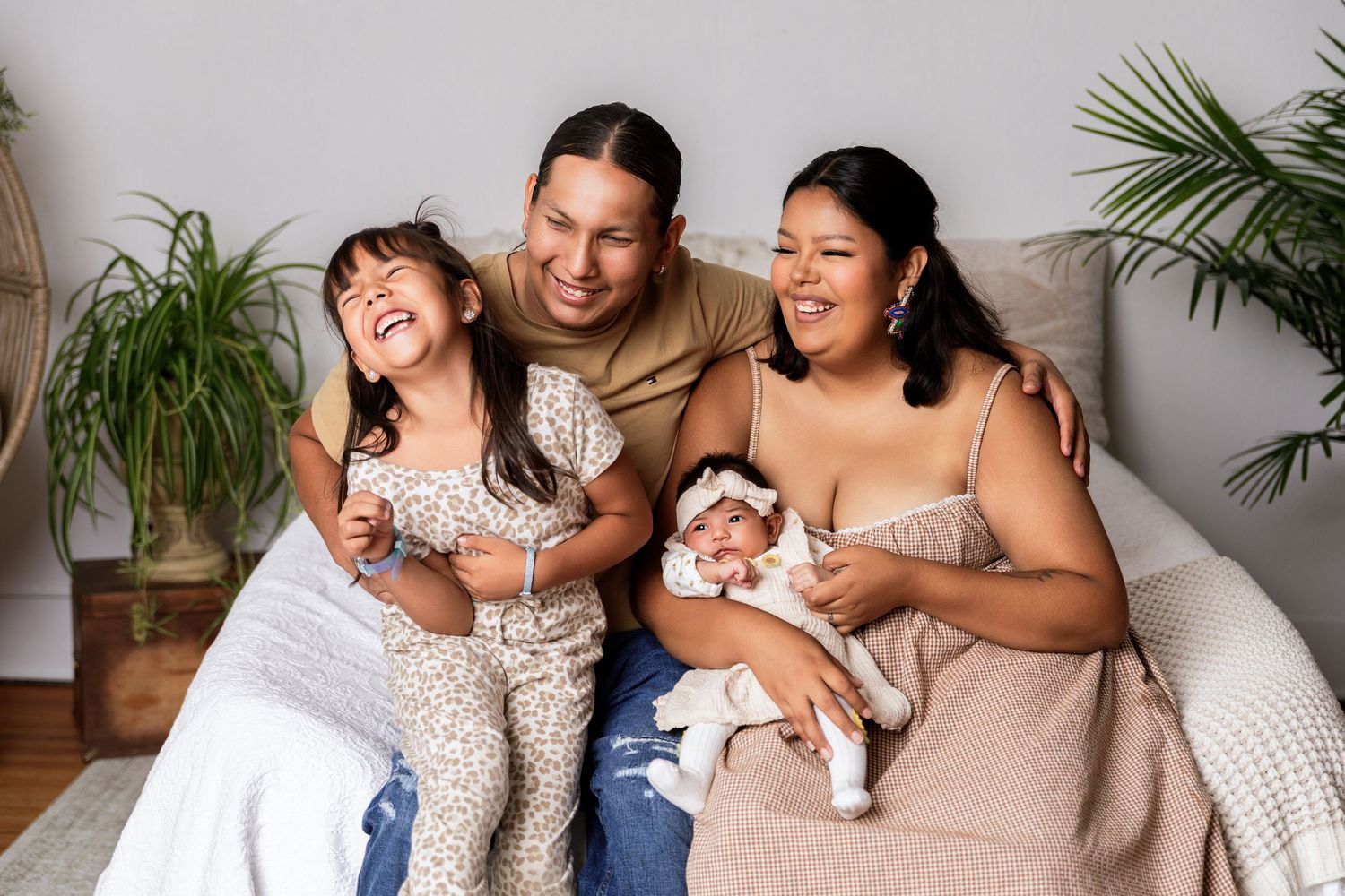Happy family of four sitting on couch, parents smiling with young daughter and newborn baby in bright living room.