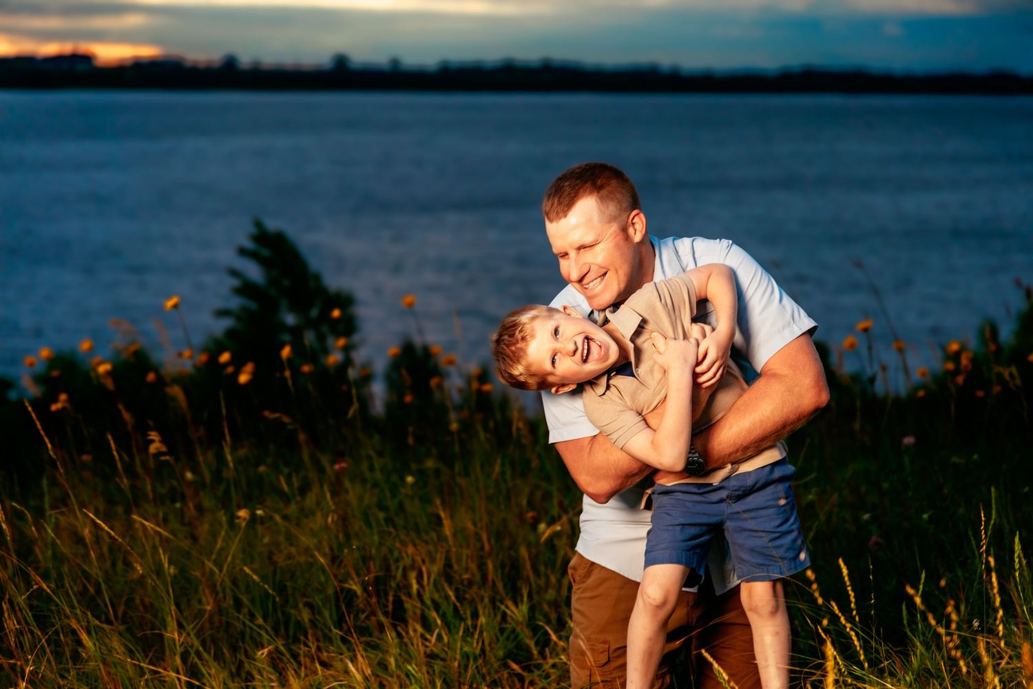 A couple embraces during a romantic sunset photo session by a waterfront with dramatic orange and blue skies.