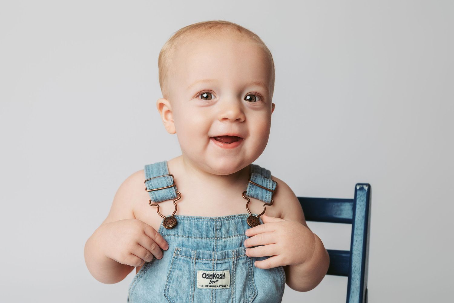Happy baby wearing light blue overalls sits in wooden chair while smiling at the camera.