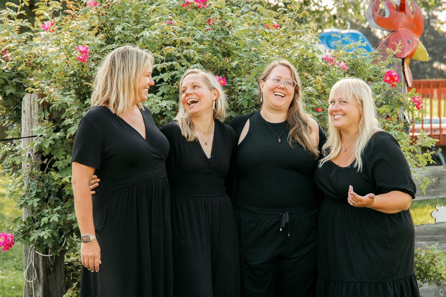 A group of people in black outfits laughing and socializing together outdoors near flowering bushes.