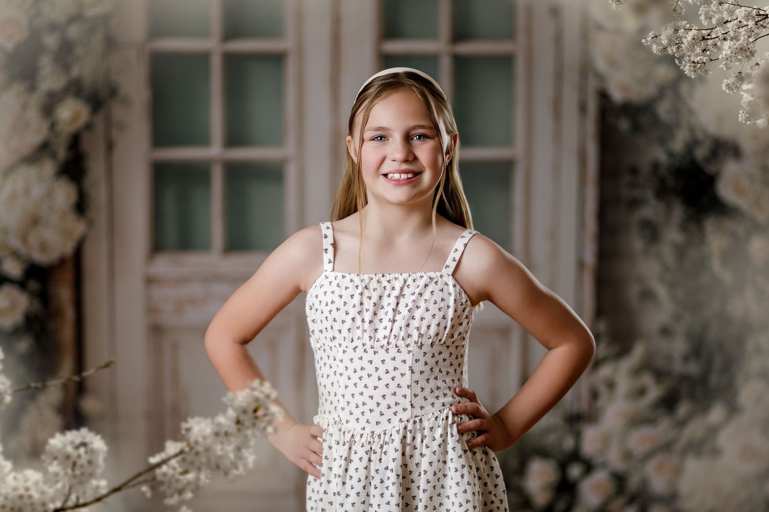Young girl in white floral sundress poses confidently with hands on hips against elegant floral backdrop.