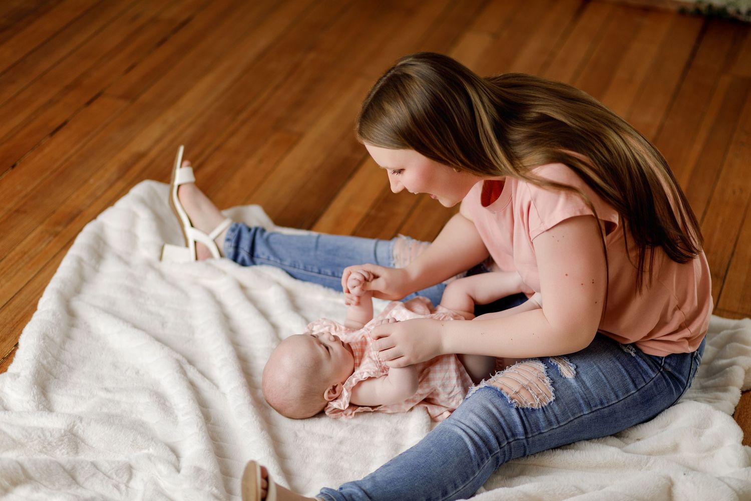 Young girl sitting on a blanket playing with a newborn baby on a hardwood floor.