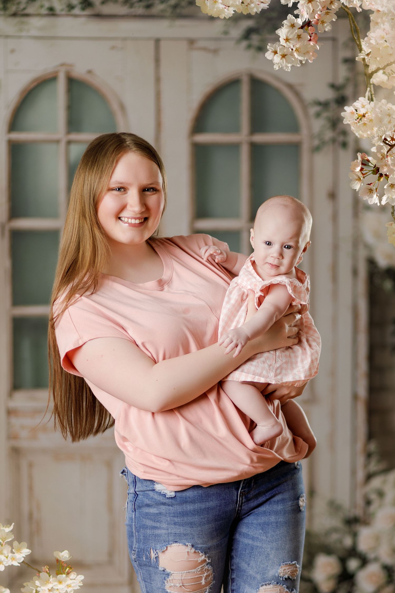 Smiling mother in pink shirt holds baby girl in pink dress before vintage floral studio backdrop.
