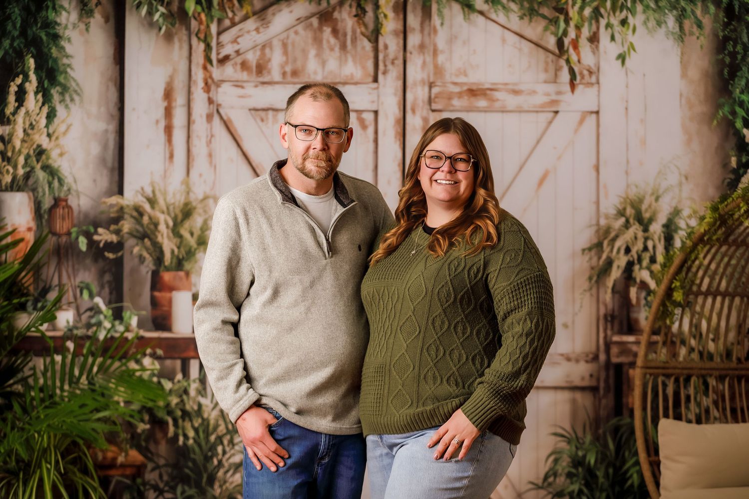 Smiling couple posing together in front of rustic barn door with green plant backdrop.