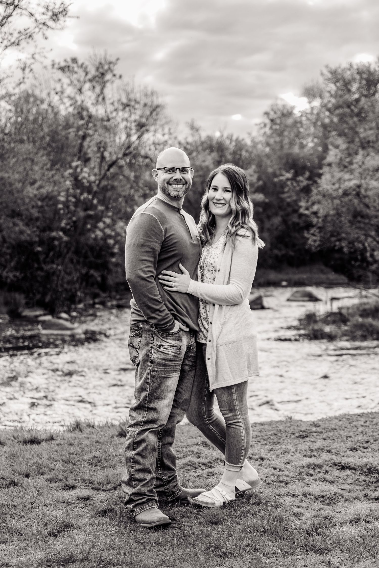 Black and white photo of a smiling couple embracing outdoors near a pond surrounded by trees.