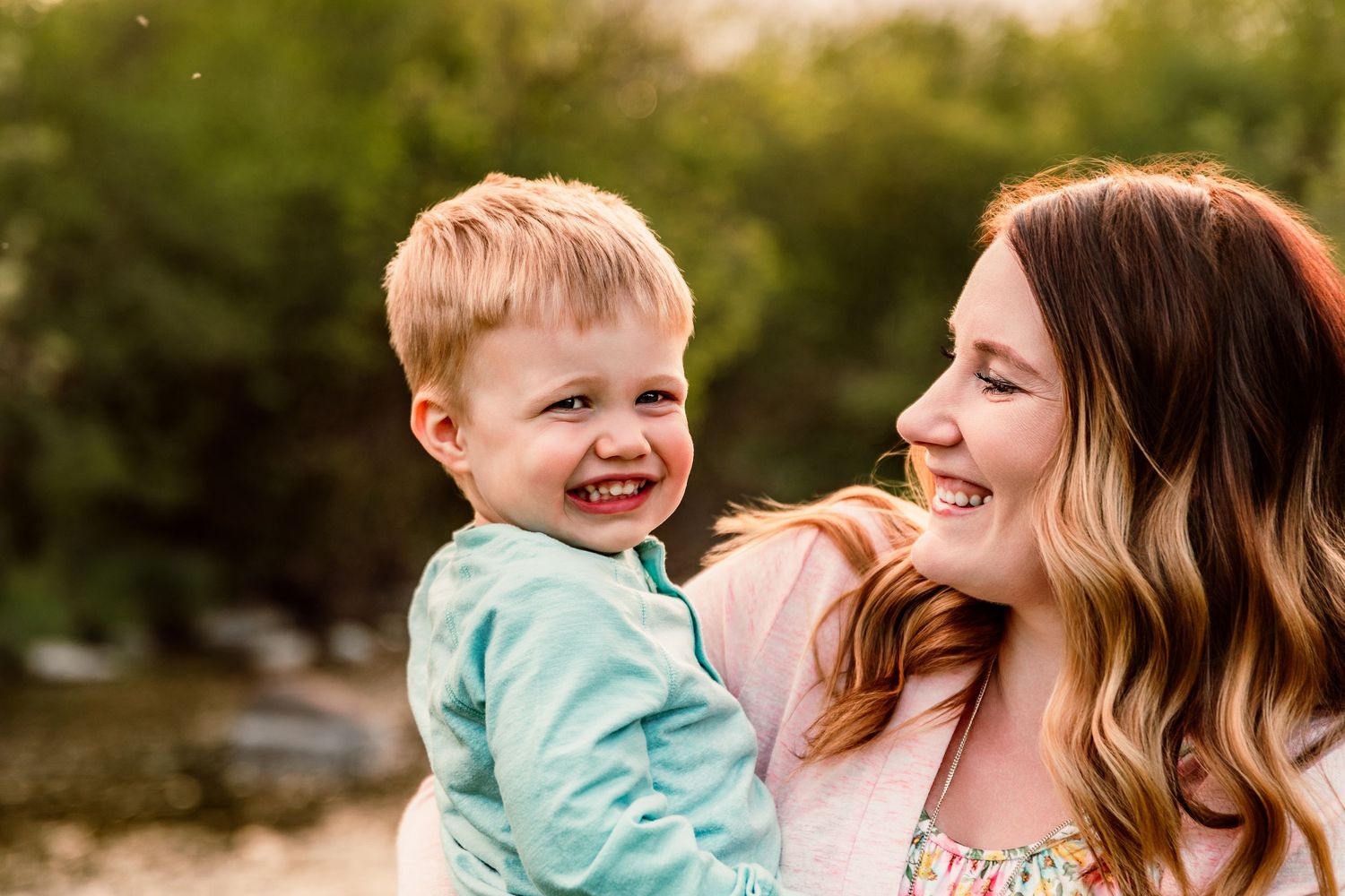 A joyful mother holds her laughing blond toddler boy outdoors, both smiling in a warm, natural setting.