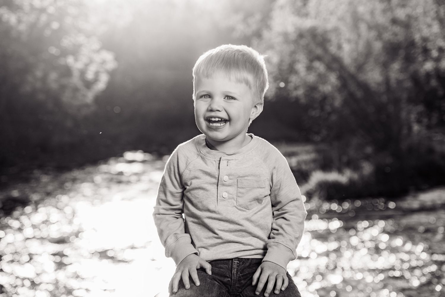 Black and white portrait of a laughing toddler boy outdoors near a sunlit stream.