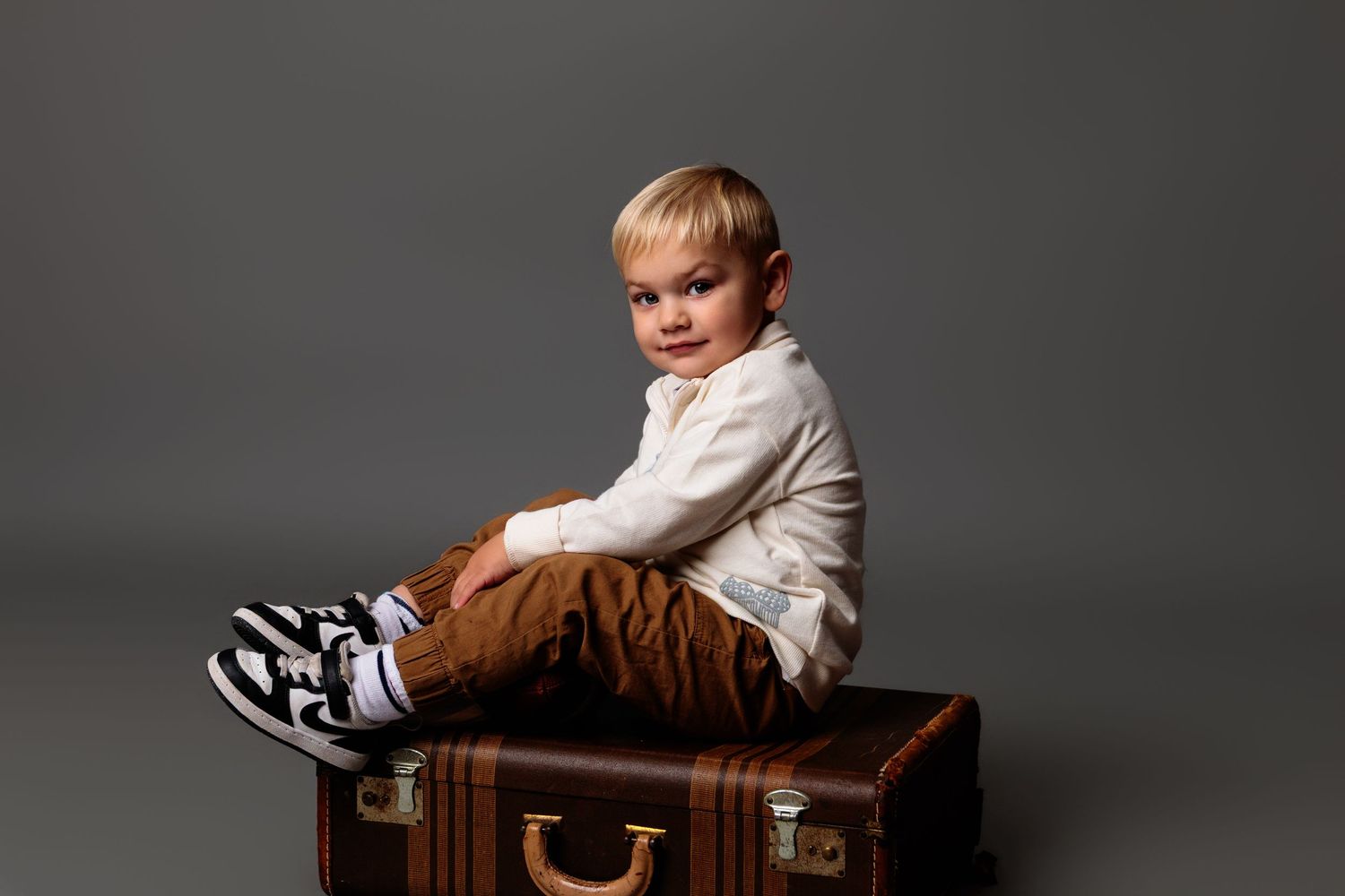 Baby sits on brown leather chair in studio against dark gray backdrop.
