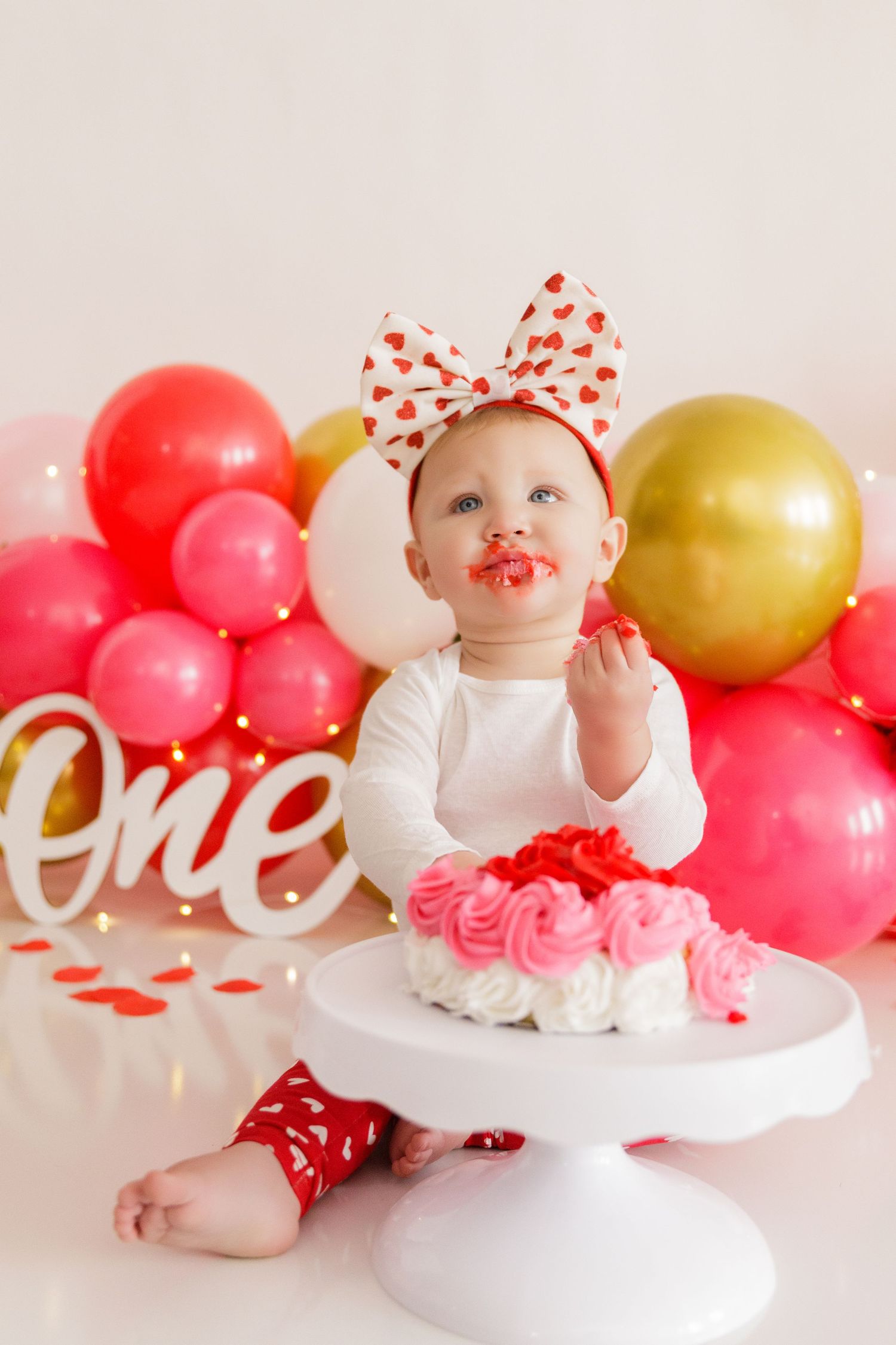 Happy baby wearing heart print bow poses with pink cake and festive balloons during Valentine's themed photoshoot.