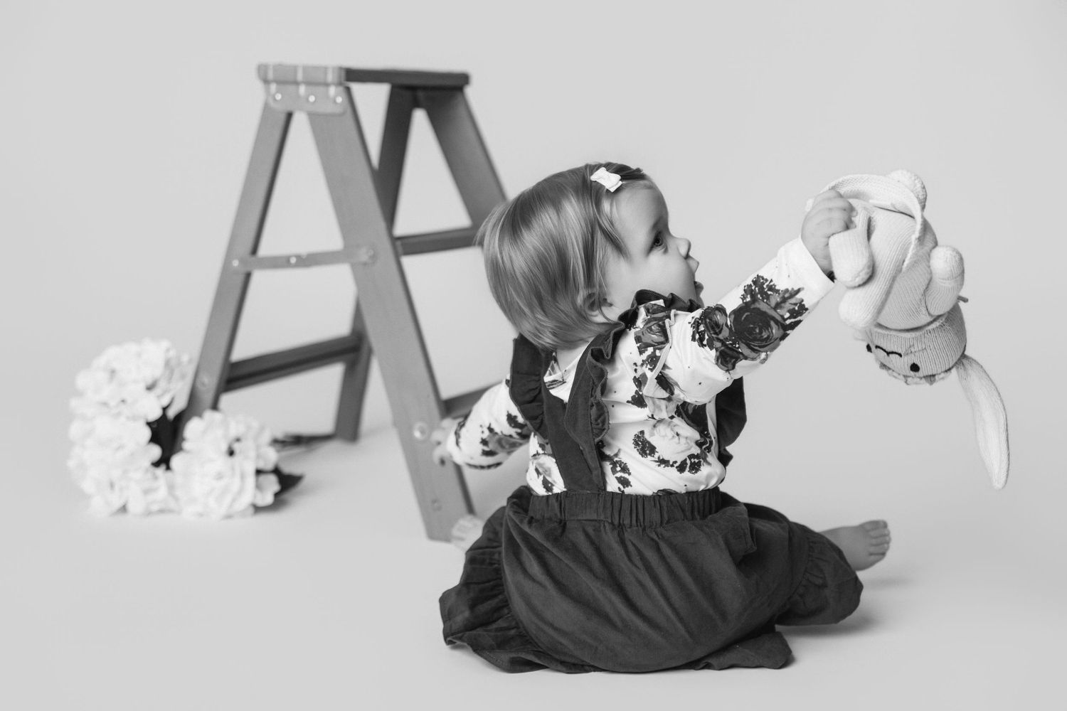 Baby in floral dress playing with stuffed toys next to wooden ladder in black and white photo.