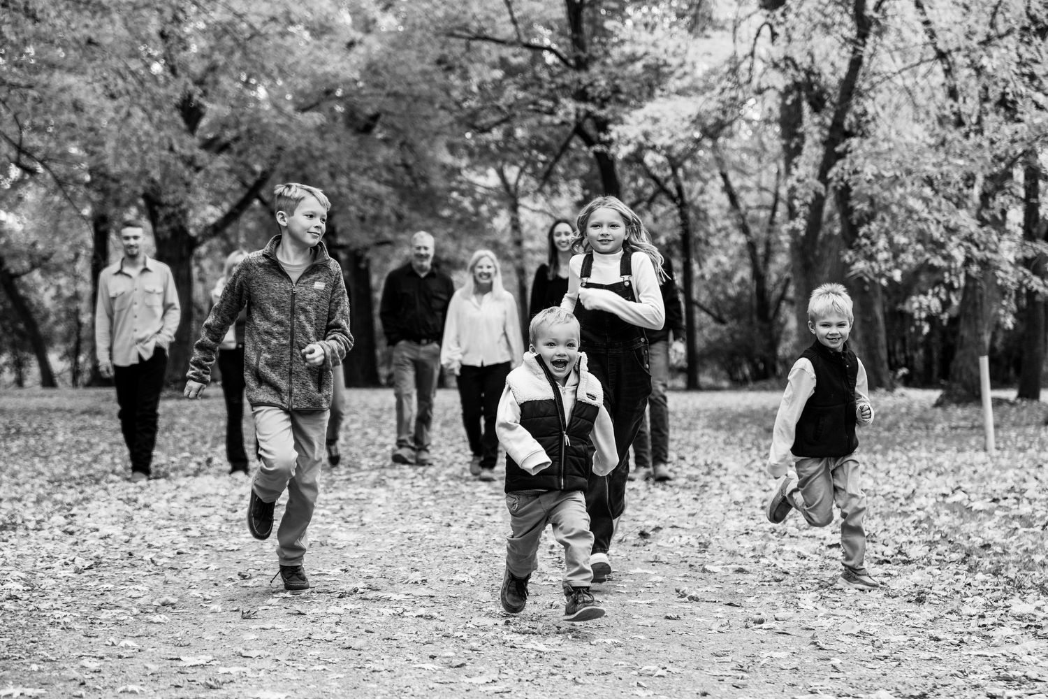 Black and white photo of people running playfully through fallen leaves in a park setting.