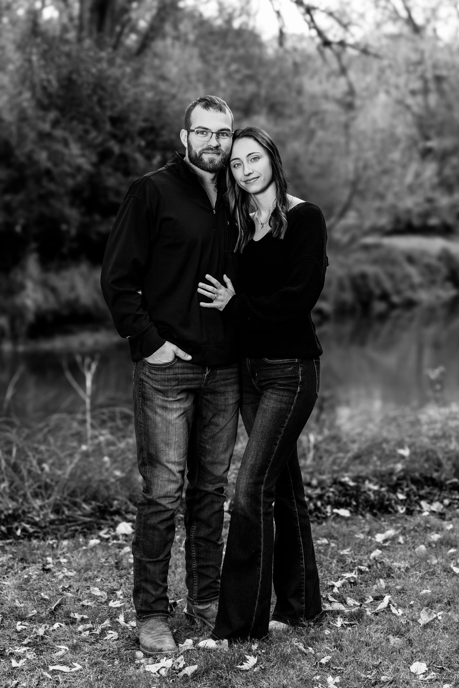 A black and white portrait of a couple embracing outdoors among fallen leaves.