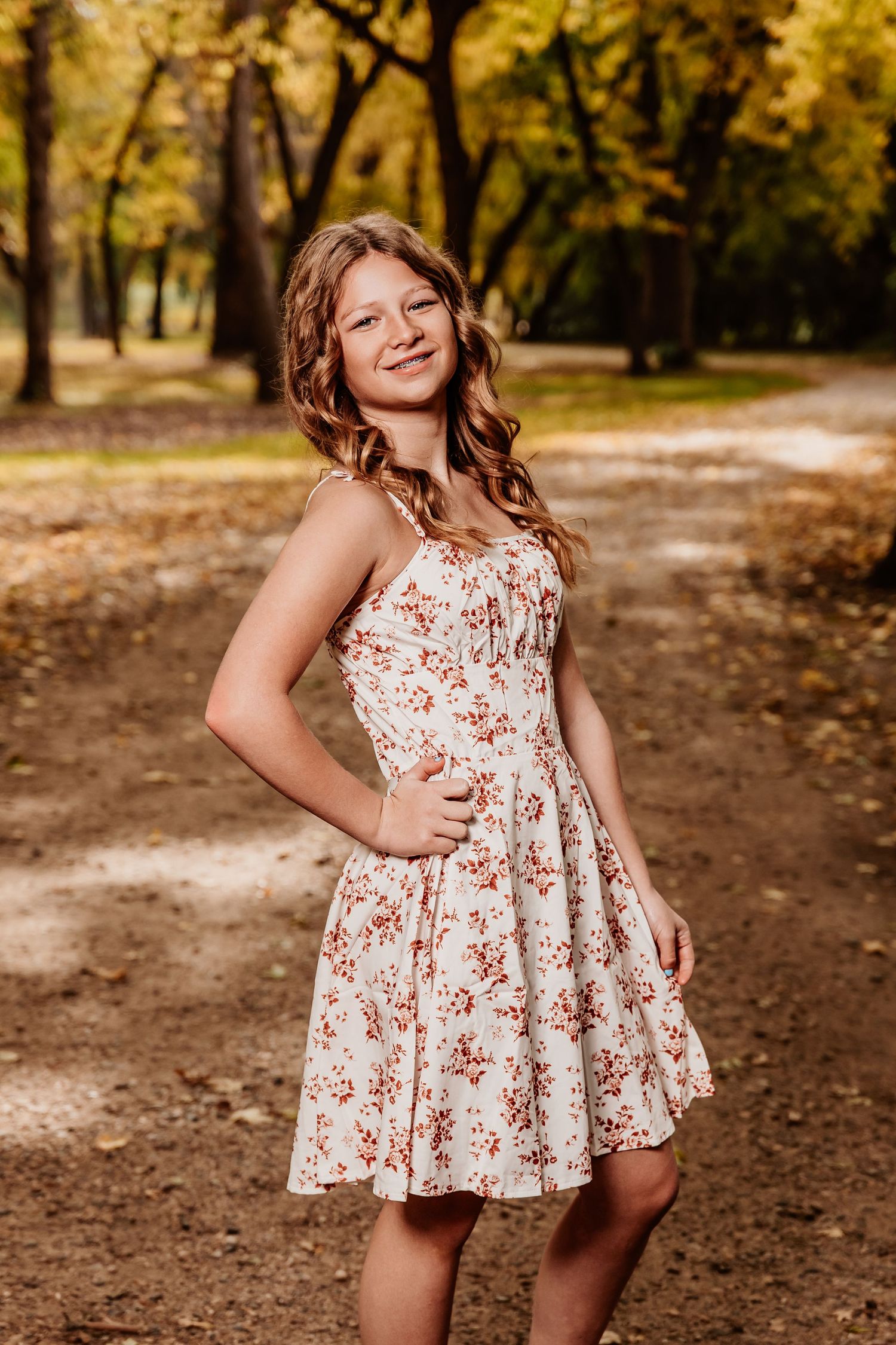 Person in a floral dress twirls playfully on a leaf-covered path in an autumn forest setting.