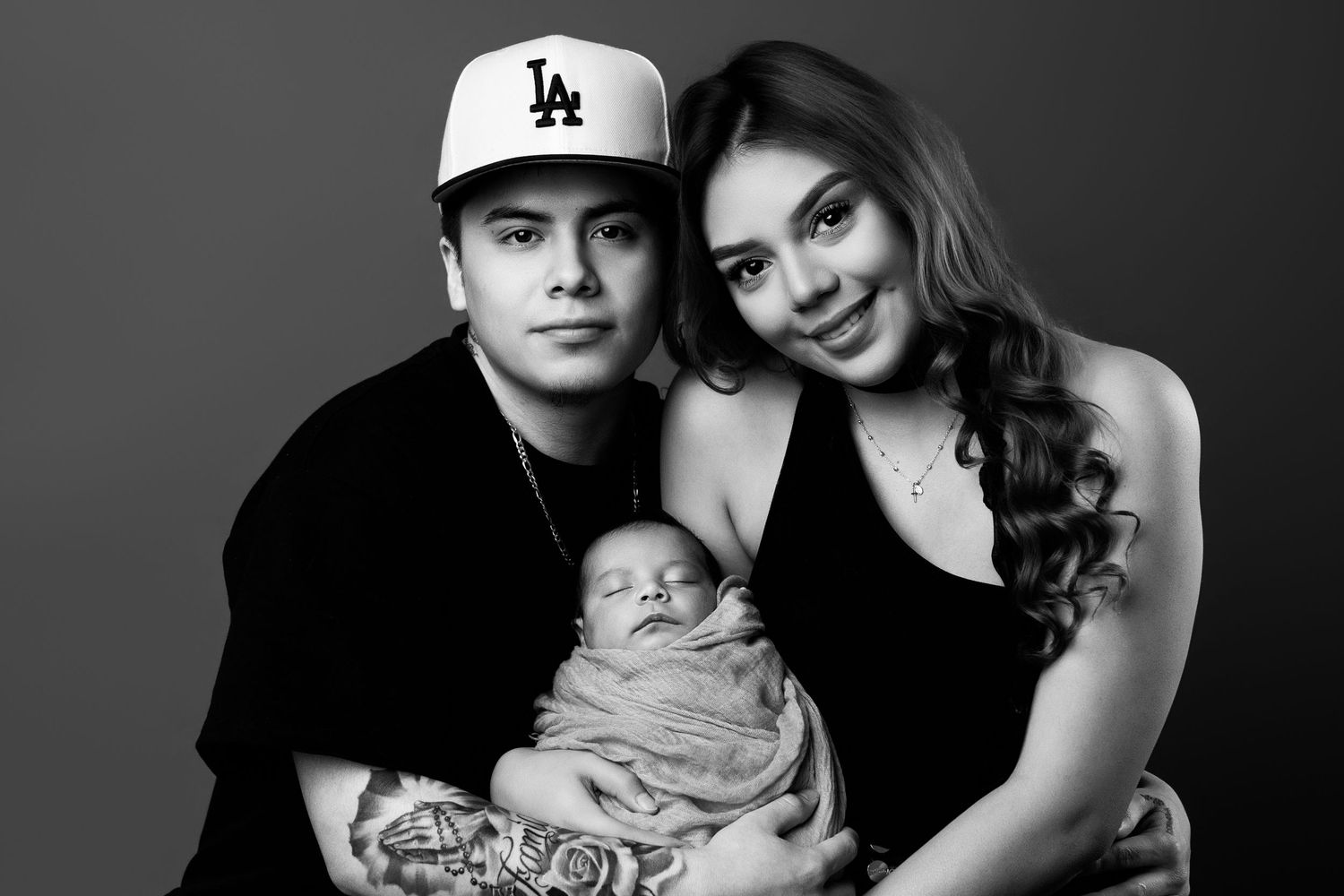 Black and white studio portrait of young couple holding their swaddled newborn baby.