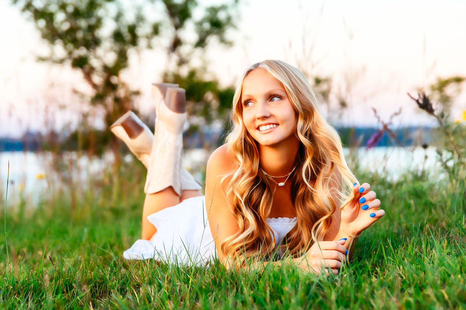 A person with wavy blonde hair lies in green grass at sunset, wearing an orange sweater while relaxing outdoors.