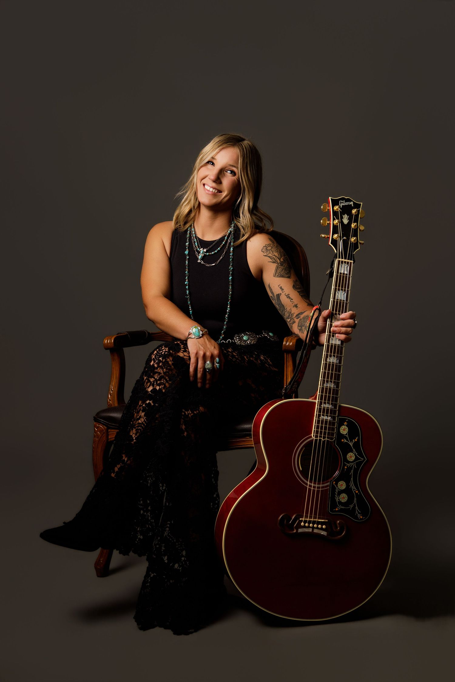 Blonde female musician in black outfit sitting with red Gibson acoustic guitar in studio.