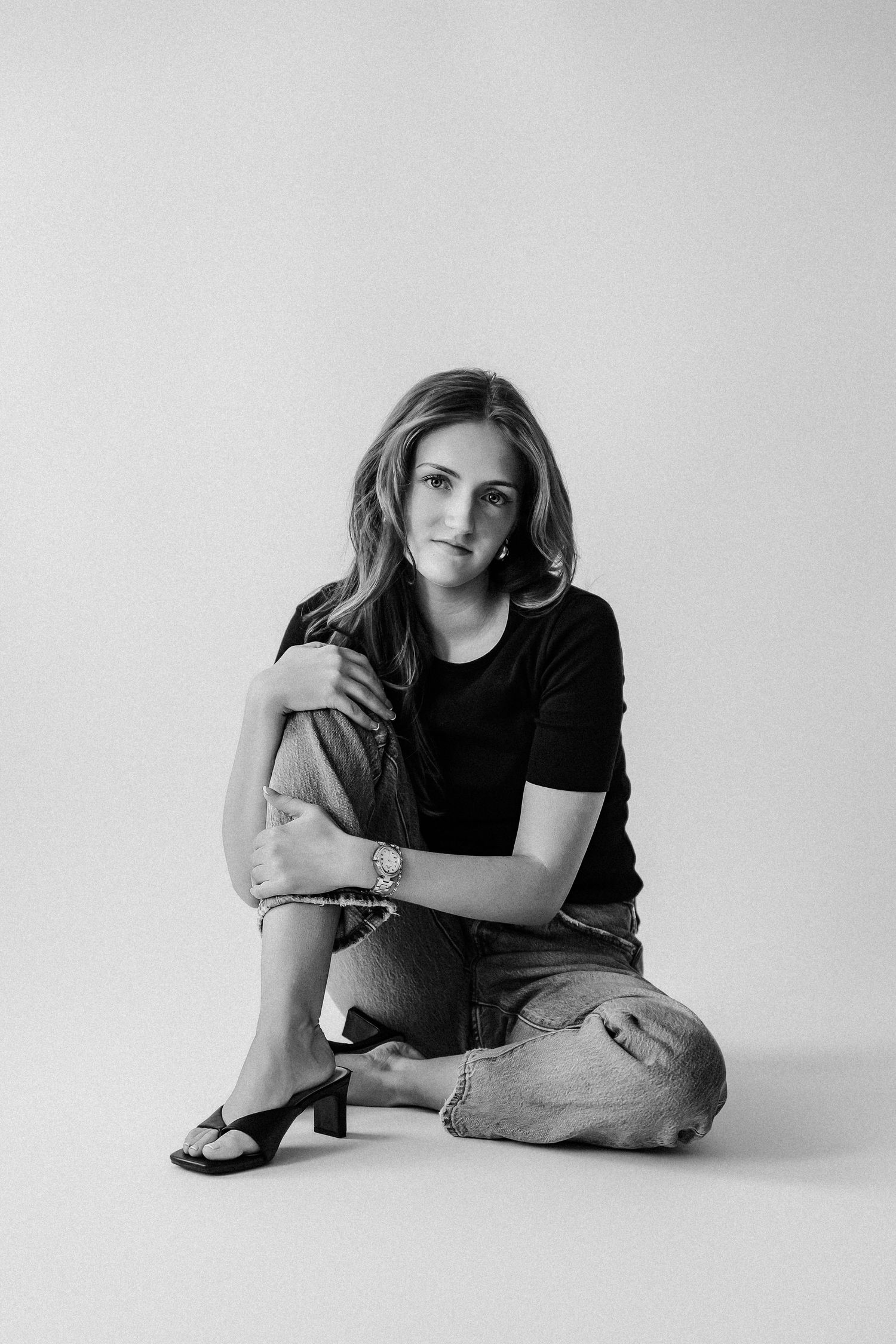 Woman in black tee and jeans sits cross-legged on floor in black and white studio portrait.