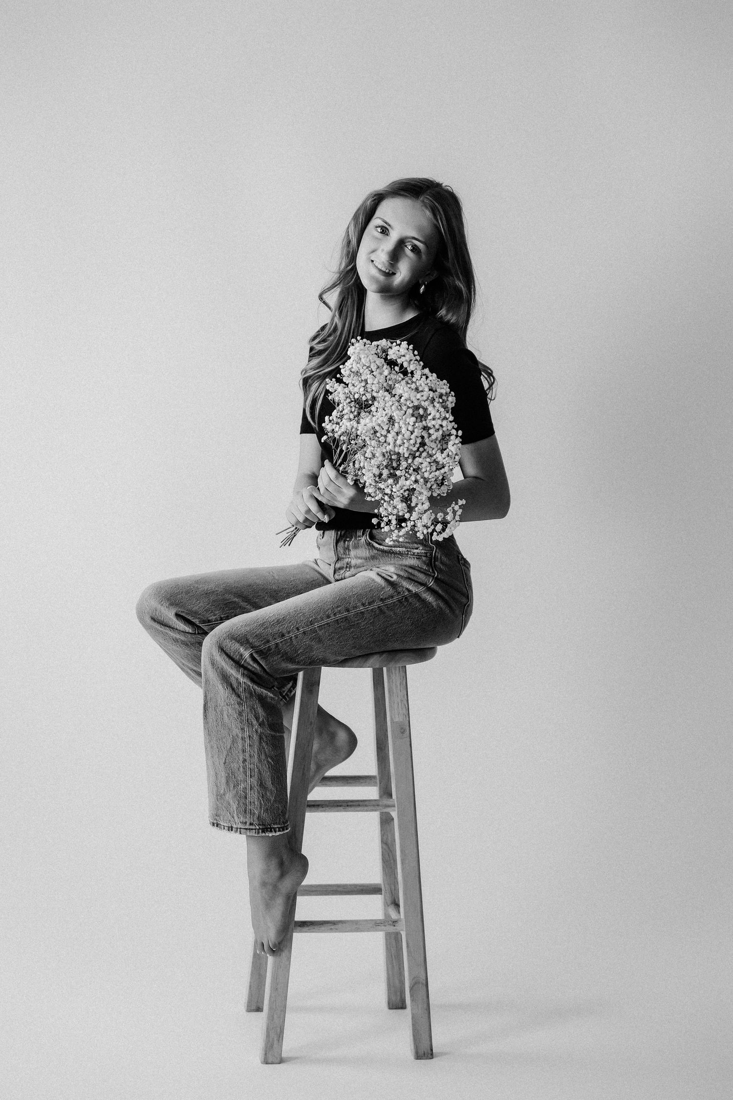 Young girl sitting on wooden stool holding white wildflowers, black and white studio portrait.