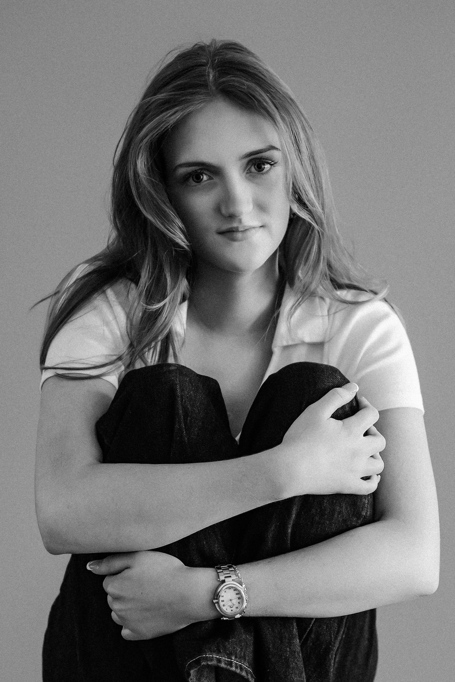 Black and white portrait of young woman in white polo hugging dark denim jacket, wearing a watch.