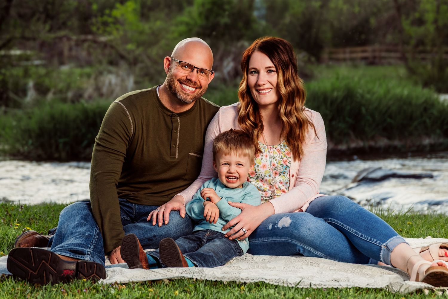 Happy family of three sitting on a blanket outdoors near a river, smiling couple with young toddler child.