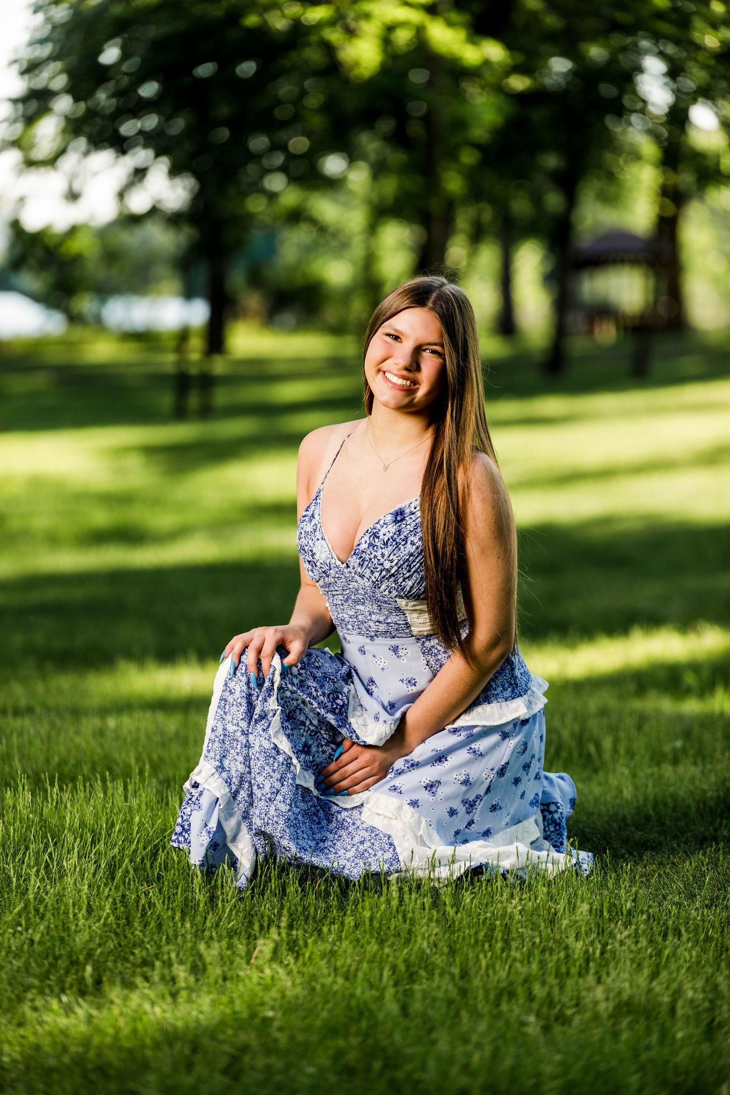 Smiling teenage girl in a blue floral dress sitting gracefully on green grass in a sunny park.